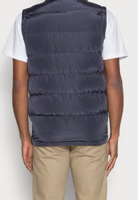 Marinblå puffväst gjord av slät, glänsande tyg, med ett horisontellt quiltat mönster och en uppstående krage, bärs över en vit skjorta.