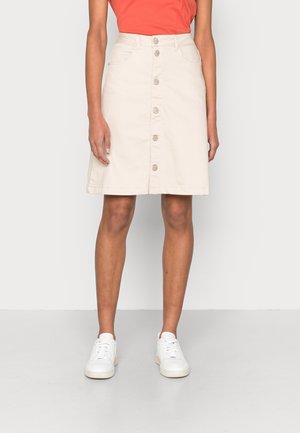 Jupe beige longueur genou avec boutons sur le devant, portée avec des baskets blanches et un haut corail, montrée de la mi-cuisse vers le bas.