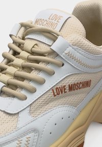 Κοντινή λήψη μπεζ και λευκού αθλητικού παπουτσιού Love Moschino με υφασμάτινη υφή, χοντρές μπεζ κορδόνια και το όνομα της μάρκας στη γλώσσα και στην πλευρά.