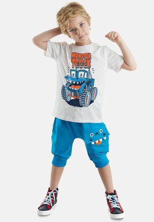 Weißes T-Shirt mit grafischem Druck eines blauen Monstertrucks. Blaues Shorts mit einem verspielten Taschendesign, das Zähne zeigt. Rote und schwarze High-Top Turnschuhe.