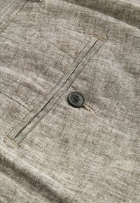 Camisa de lino marrón con un bolsillo en el pecho, un detalle de botón negro y una tela texturizada con sutiles variaciones en el tono.