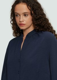 Navyblaue Bluse aus strukturiertem Stoff, mit hohem Kragen und Knopfverschluss. Verfügt über dezente Falten entlang des Ausschnitts.