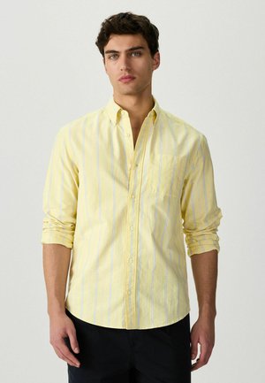 Młody mężczyzna ubrany w jasnożółtą, długorękawową koszulę na guziki z pionowymi białymi i niebieskimi paskami, z podwiniętymi rękawami, stojący na jednolitym tle.
