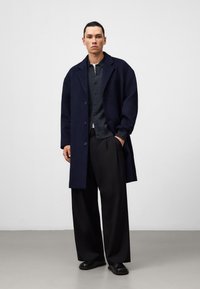 Marineblauer Wollmantel über einem dunklen, strukturierten Hemd, kombiniert mit weiten schwarzen Hosen und schwarzen Schuhen.