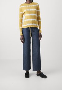 Żółty sweter prążkowany z białymi paskami, w połączeniu z ciemnoniebieskimi spodniami typu wide-leg i czarnymi butami slip-on. Sweter ma regulowany sznurek na dole.