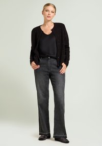 Cardigan nero con maglia texturizzata e sottili decorazioni, abbinato a jeans scuri a zampa. Top nero sotto, scarpe nere, sfondo neutro.