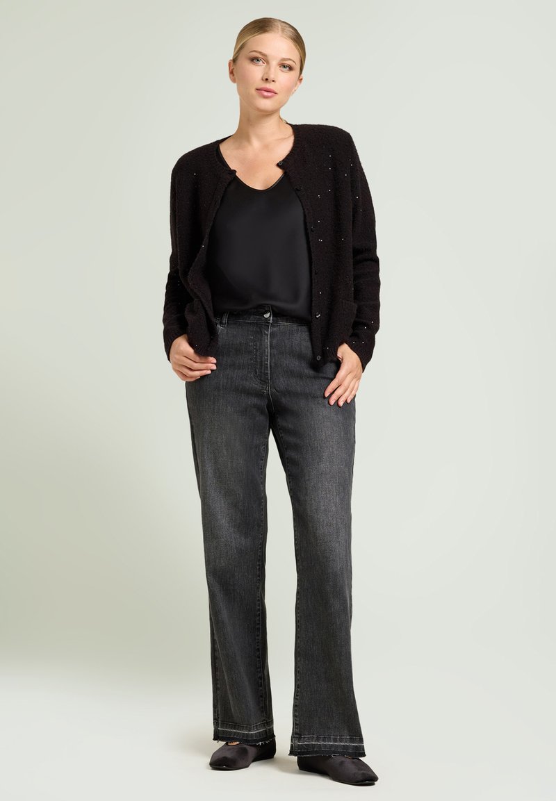 Cardigan nero con maglia texturizzata e sottili decorazioni, abbinato a jeans scuri a zampa. Top nero sotto, scarpe nere, sfondo neutro.