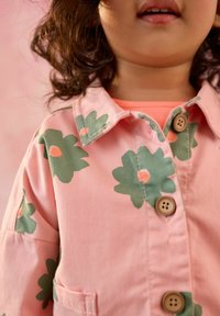 Rosa jacka med gröna blommönster och persikofärgade detaljer. Har en krage, två knappar och fickor. Tyget är mjukt och texturerat.