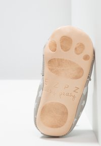 Zapato suave gris con suela blanda, patrón de estrellas plateadas, parte superior de cuero y fondo de goma color marrón claro con un diseño de huella de pata y nombre de la marca.