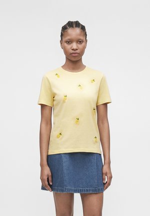 Kvinde med flettet hår iført en lys gul t-shirt med små ananas og et blåt denimskørt, stående mod en ensartet lys baggrund.