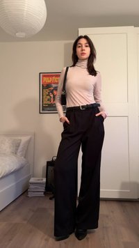 Εφαρμοστό, ανοιχτό ροζ πουλόβερ με ζιβάγκο, μαύρο παντελόνι με φαρδιά μπατζάκια, ψηλόμεσο, με πιέτες, μαύρη ζώνη και μαύρες γόβες. Απλός διακοσμητικός φόντος.