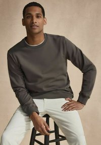 Mörkgrå sweatshirt med rund halsringning, ribbad mudd och en slät yta, i kombination med ljusfärgade byxor, sittande på en pall.