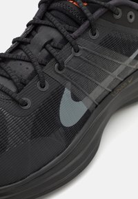 Svart atletiskt sko med texturerat meshtyg, förstärkta snörhål, en ljusgrå logotyp och orange detaljer längs den övre delen.