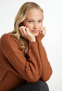 Maglione lavorato a maglia color ruggine con polsini a coste, caratterizzato da un motivo testurizzato. Modella in posa con le mani vicino al viso, su uno sfondo bianco.