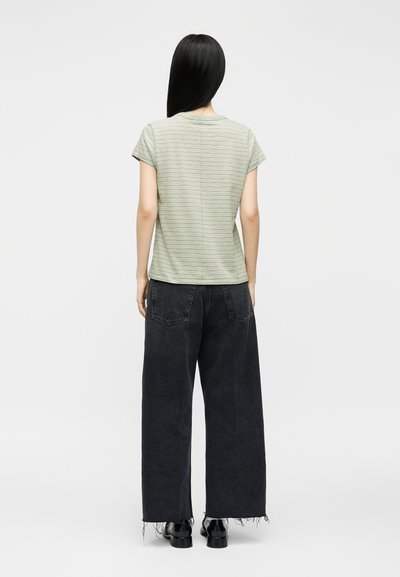 Γυναίκα με μακριά ίσια μαύρα μαλλιά φοράει ανοιχτό πράσινο ριγέ κοντομάνικο πουκάμισο, φαρδιά μαύρα τζιν με ξεφτισμένα τελειώματα και μαύρα παπούτσια, όρθια.