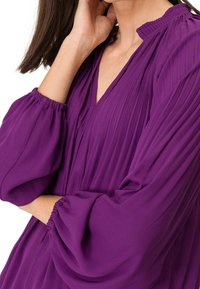 Valkoinen pleatattu paita, jossa on V-pääntie ja kerätty hihansuu. Kangas on kevyt, mikä luo virtaavan siluetin pehmeällä tekstuurilla.