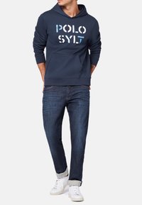 Marinblå huvtröja med vit och ljusblå text "POLO SYLT", känguruficka, kombinerad med mörka jeans och vita sneakers.