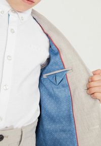 Giacca di lino grigia chiara con foderatura a fantasia blu e rifiniture rosse. Camicia bianca con colletto button-down sottostante, che mostra un branding sottile.