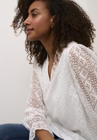 White blouse featuring intricate diamond patterns and lace detailing, with long, puffed sleeves and a relaxed fit. Worn with denim.