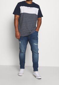 T-shirt en coton coloré en blocs bleu marine et gris avec une bande blanche en haut, associé à un jean bleu déchiré et des baskets blanches.