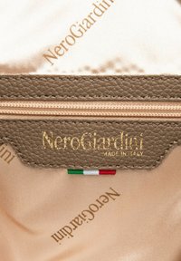 Etiqueta de la marca NeroGiardini en oro sobre cuero marrón texturizado con fondo de satén crema. Presenta los colores de la bandera de Italia y el texto "HECHO EN ITALIA".