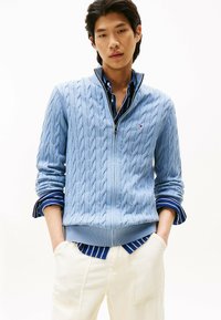 Helles blaues, kabelgestricktes Sweatshirt mit Reißverschlusskragen, über einem gestreiften blauen Button-up-Hemd getragen. Das Model trägt cremefarbene Hosen.