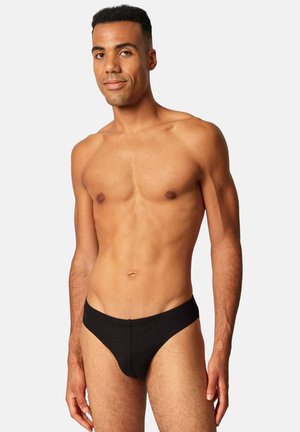 Mann steht in schwarzer Unterhose, schaut frontal und hat einen neutralen Gesichtsausdruck vor einem einfarbigen hellgrauen Hintergrund.