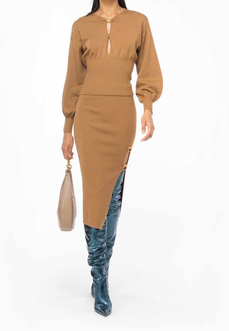 Femme portant une robe en maille marron avec un décolleté en trou de serrure et une fente latérale, des bottes métalliques bleues jusqu'aux genoux, et tenant un sac à main beige.