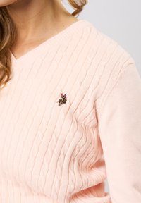 Světle růžový svetr s copánkovým vzorem, výstřihem do V a texturovaným vzorem, na levé straně s malou vyšívanou broží ve tvaru květin.