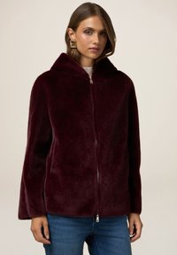 Giacca in finta pelliccia bordeaux con cappuccio, dotata di chiusura frontale con zip, vestibilità comoda e texture morbida, abbinata a un top in maglia e jeans blu.