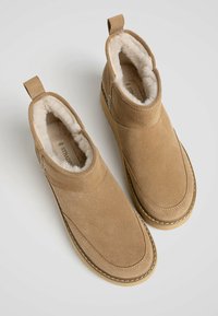 Bottines en daim beige avec une doublure en fourrure douce, bout arrondi et un détail cousu le long de la semelle. Onglets à l'arrière pour un enfilage facile.