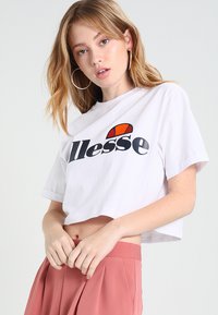 Weißes, kurzes T-Shirt mit "ellesse"-Logo in Schwarz und Rot. Es hat kurze Ärmel und eine lässige Passform und wird mit rosa hochgeschnittenen Hosen kombiniert.
