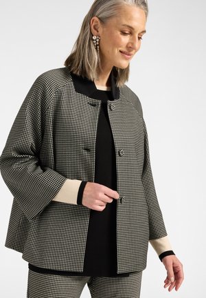 Frau trägt eine schwarz-weiß klein karierte Jacke und passende Hosen, mit Perlen-Ohrringen und einem schwarzen Top darunter.