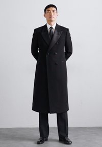 Melnā virsjakā ar dubultu pogu dizainu, satīna atlokiem, pieguļošu piegriezumu un gludu tekstūru, kopā ar oficiālām biksēm un spīdīgām kurpēm.