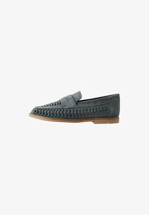 Mocassino in suede grigio con dettagli intrecciati sui lati e sul collo del piede, suola piatta color cuoio, stile slip-on, visto di lato su sfondo bianco.