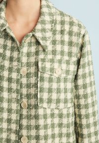Green and cream checkered button-up shirt made of textured fabric, featuring a collar, pocket, and rounded buttons.