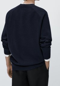 Pull en maille bleu marine avec poignets et ourlet côtelés. Présente un col rond et des manches raglan. Porté sur une chemise blanche.