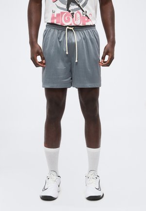 Persona que lleva puestos unos pantalones cortos de malla grises con cordón, calcetines deportivos blancos, zapatillas blancas y una camiseta gráfica con el texto "Hoops" visible.