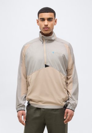 Man draagt een beige en lichtgrijs vest met ritssluiting en elastische manchetten, en groene broek, staand tegen een effen witte achtergrond.