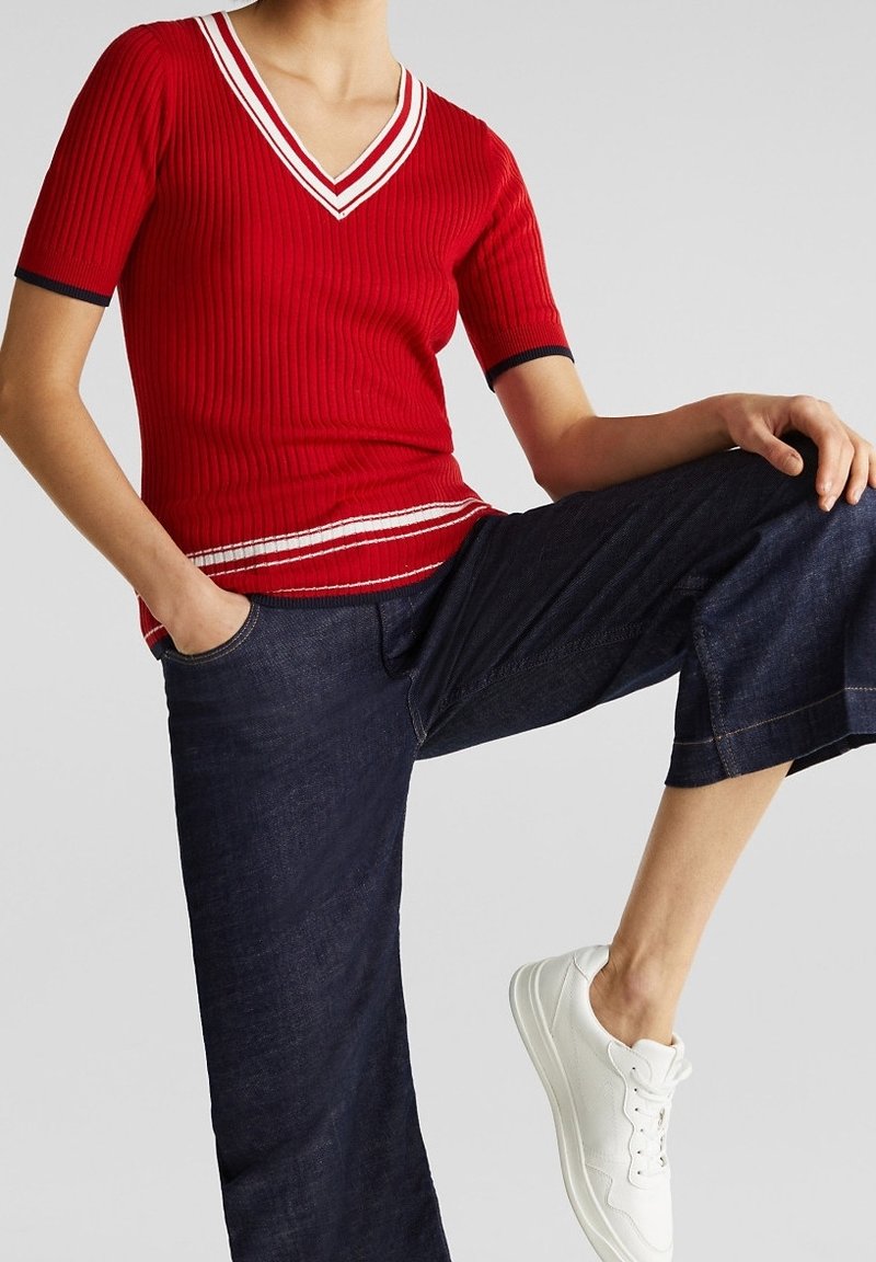 Top corto de rayas rojas con cuello en V y rayas blancas y marinas en el escote y el dobladillo. Combinado con unos pantalones de mezclilla de pierna ancha de color azul oscuro y zapatillas blancas.