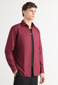 Bordeaux langærmet skjorte med knaplukning foran, sorte knapper og spredt krave båret over en sort t-shirt. Lavet af glat stof.
