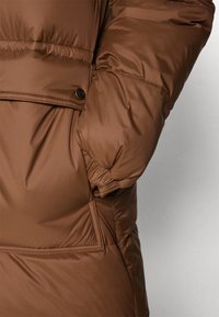 Chaqueta puffer aislante marrón con un acabado texturizado, que presenta un bolsillo lateral con cierre de botón y puños elásticos para mayor calidez.