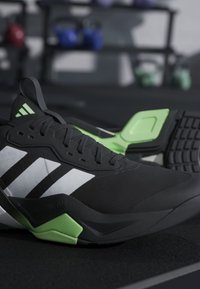 Chaussure de sport Adidas noire et verte avec des bandes blanches posée sur un sol noir dans une salle de sport avec des poids flous en arrière-plan.