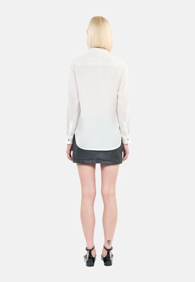 Langemouwige, semi-doorzichtige witte blouse met knoopmanchetten, gecombineerd met een zwarte leren minirok. Soepele textuur, gebogen zoom en getailleerd ontwerp.