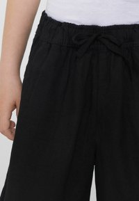 Černé, strukturované kalhoty s elastickým pasem a šňůrkou. Silueta je volná, sahající do středu lýtek.