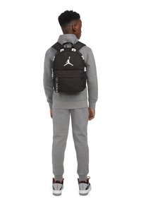 Črn nahrbtnik z belim Jordan logotipom in odločno napisano ime. Ima dva nastavljiva traku in žep z zadrgo, poenostavljen dizajn in teksturirano tkanino.