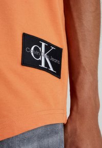 Oranžový lem trička s černou nášivkou loga Calvin Klein Jeans, nošeno s modrými džíny, a viditelný předloktí osoby.