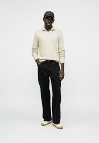 Pullover lavorato a maglia crema con zip a metà, jeans neri e sneakers beige. Il cappellino è nero e lui sta in piedi con le mani in tasca su uno sfondo neutro.