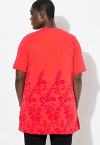 Haut rouge à manches courtes avec un motif floral dans une teinte rouge plus foncée à l'ourlet. Fabriqué en tissu lisse avec des fentes latérales et une coupe décontractée.