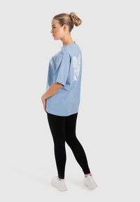 Lys blå oversized t-shirt med korte ærmer, med et hvidt grafisk design på ryggen. Bæres med sorte leggings og hvide sneakers.